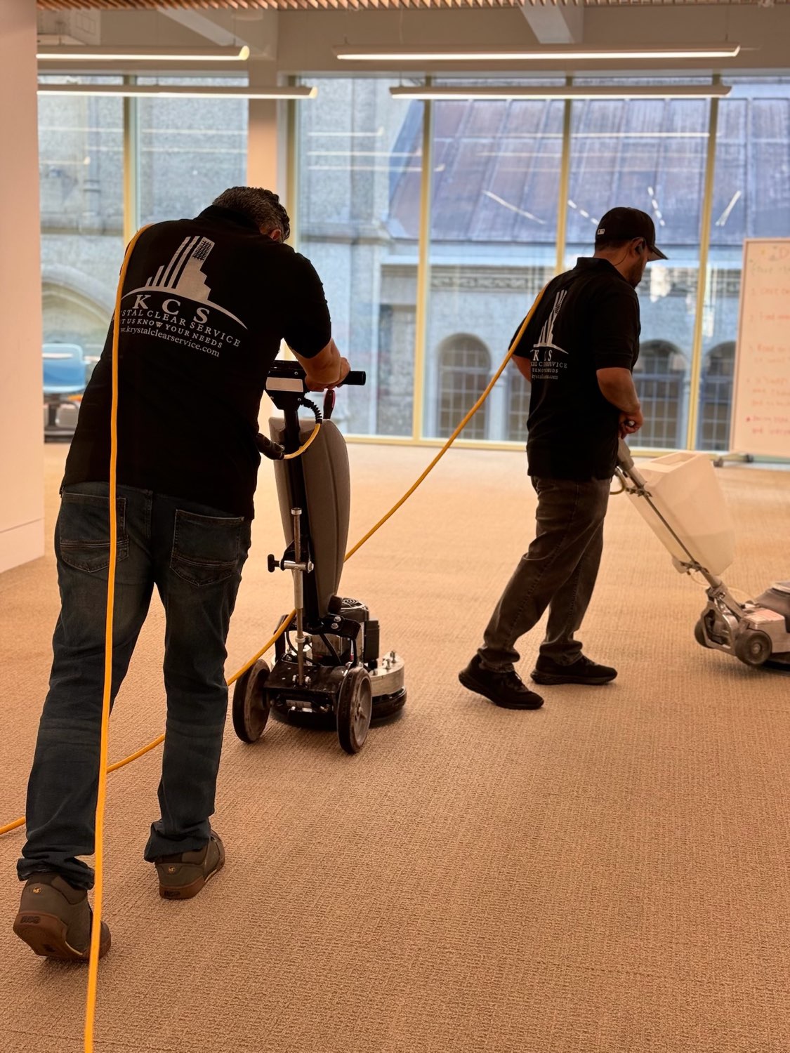 Two KCS crew members in branded shirts running carpet machines in a commercial building